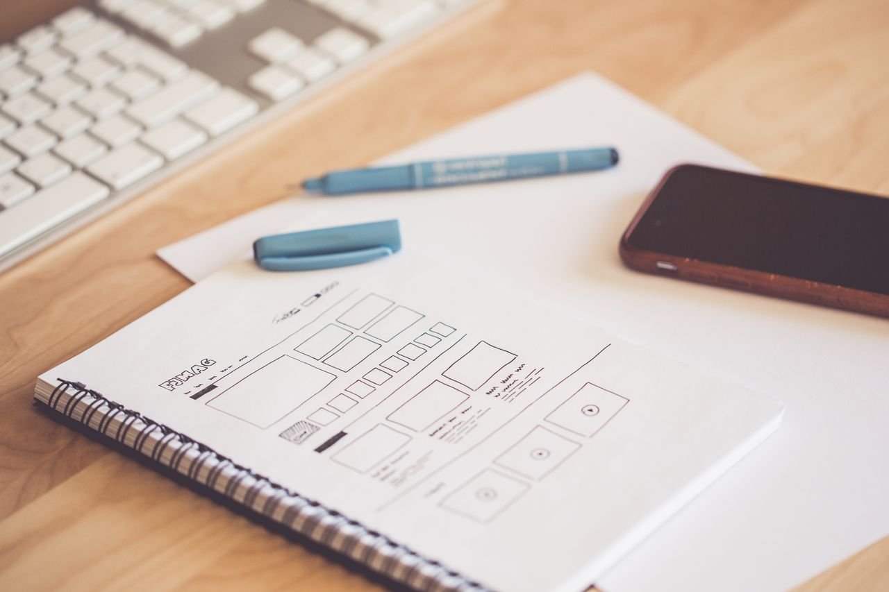 Mastering the First Impression: Your intriguing post title goes here A neat workspace featuring a sketchpad with wireframes, a smartphone, and a keyboard on a wooden desk.